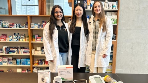 Three people wearing white lab coats stand behind a table displaying home healthcare devices, including a blood pressure monitor, glucose meter, inhalers, and medical packaging. Shelves stocked with boxed pharmacy products line the background in a bright retail or clinical setting.