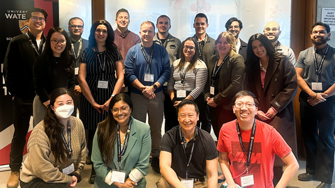 Group photo of approximately 18 adults wearing conference lanyards, arranged in two rows inside a modern indoor meeting space. The individuals are dressed in business-casual attire and stand or kneel in front of a light-colored wall with partial signage visible, including University of Waterloo branding and a sign that reads “Quiet Please Meeting in Progress.” Lighting is bright and even, and the image appears to document a professional gathering or workshop.