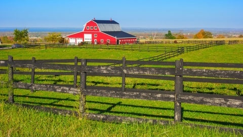 A bright red barn with a dark roof sits beyond green fields and black wooden fences under a clear blue sky, with rolling countryside in the distance. 