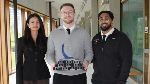 Three students pose in a campus hallway, with one holding a multi‑plate award trophy.
