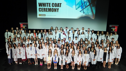 Large group of University of Waterloo pharmacy students in white coats posed on stage under a “White Coat Ceremony” banner, with school banners on either side.