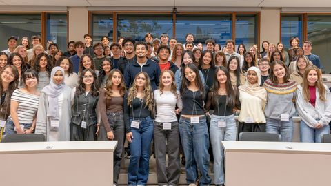 A large group of students pose together for a group photo in a bright classroom, arranged in several rows between desks and wearing name badges.