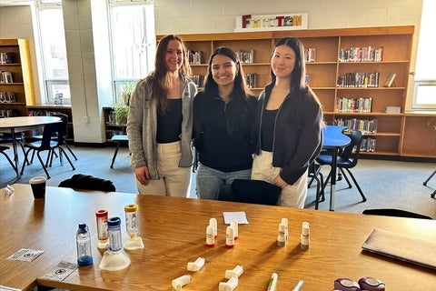 Three people wearing white lab coats stand behind a table displaying home healthcare devices, including a blood pressure monitor, glucose meter, inhalers, and medical packaging. Shelves stocked with boxed pharmacy products line the background in a bright retail or clinical setting.