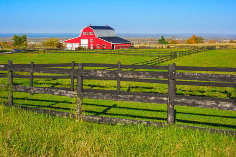 A bright red barn with a dark roof sits beyond green fields and black wooden fences under a clear blue sky, with rolling countryside in the distance. 