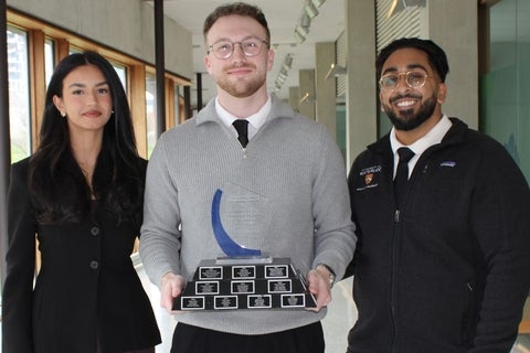 Three students pose in a campus hallway, with one holding a multi‑plate award trophy.