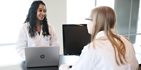 two students in white coats talking at desk