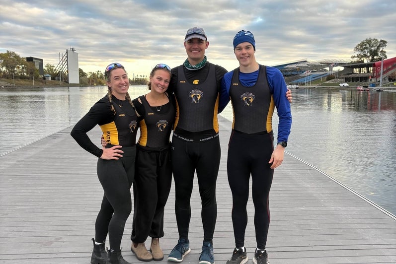Four rowing team members stand together on a wooden dock beside a calm watercourse under a cloudy sky. All are wearing black and gold Waterloo Warriors rowing uniforms, with one person in a blue long-sleeve layer and another wearing a cap. The group stands close together with arms around each other. In the background, there is a rowing basin with lanes, a starting tower, and grandstand seating along the shoreline.