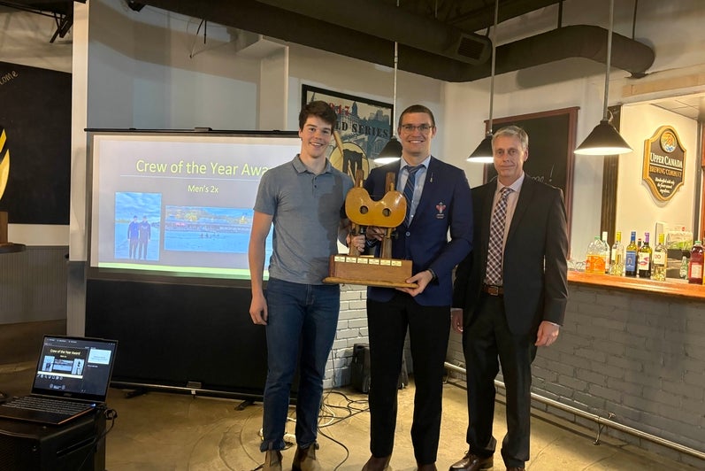 Three people stand indoors at an awards event, with one person holding a wooden trophy. A presentation screen behind them reads “Crew of the Year Award – Men’s 2x” and shows rowing photos. The setting includes a projector, a bar counter with bottles, and wall decor, under warm overhead lighting.