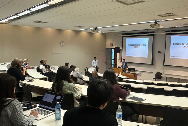 Tiered lecture hall with attendees seated at long desks using laptops and notebooks, facing a speaker at a podium. A presentation slide on screen reads “Pharmacy Research Day 2026.”