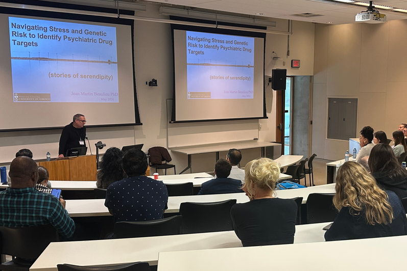 A speaker presents at the front of a lecture hall beside two large projection screens displaying a slide titled “Navigating Stress and Genetic Risk to Identify Psychiatric Drug Targets,” while attendees sit and listen.