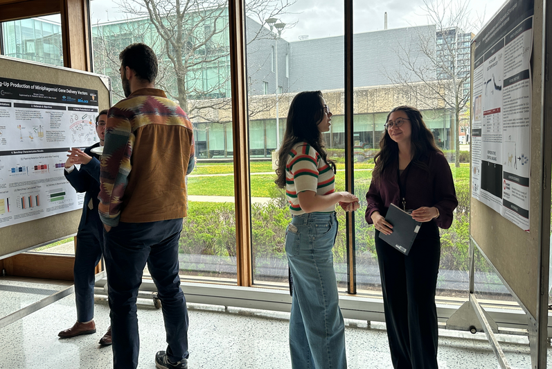 Group of attendees gathered around a research poster display, engaging in discussion and viewing charts and text on the board, with campus buildings visible through the windows in the background.