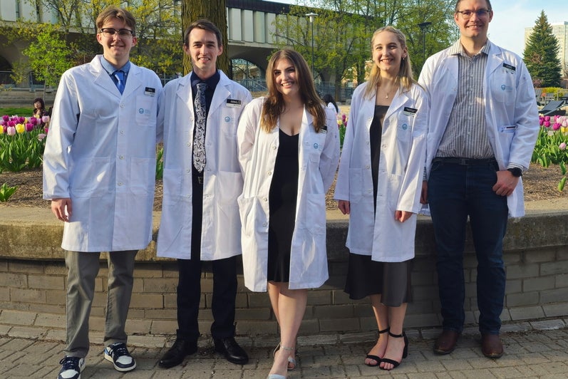 Five people wearing white lab coats stand outdoors in a landscaped campus setting with blooming tulips and leafy trees. They are positioned in a line in front of a low brick wall, with a modern building visible in the background. The group faces the camera, and some wear name tags and formal attire underneath their lab coats.