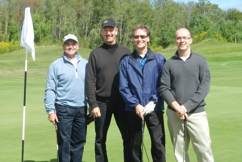 Four men posing by golf flag