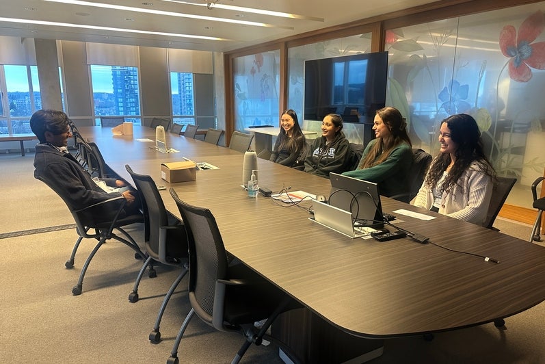 Large conference room with a long rectangular table and rolling office chairs. One adult sits slightly apart at one end of the table, while a small group of adults sit along the opposite side, all oriented toward one another during a discussion. Laptops, cables, a reusable water bottle, and a small box rest on the tabletop. The room features glass walls with floral designs, large windows showing an urban skyline, a ceiling‑mounted projector, and bright overhead lighting, suggesting a formal meeting, intervi
