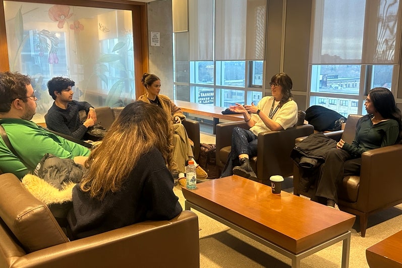 Group of adults seated in armchairs arranged in a circle around a low rectangular coffee table in a lounge-style meeting area. One participant gestures while speaking as others sit facing inward, participating in the discussion. The space has large windows with partially lowered blinds, letting in daylight and revealing nearby buildings outside. Coats, bags, notebooks, a water bottle, and takeaway coffee cups rest on chairs and the table, suggesting an informal group discussion or facilitated conversation i