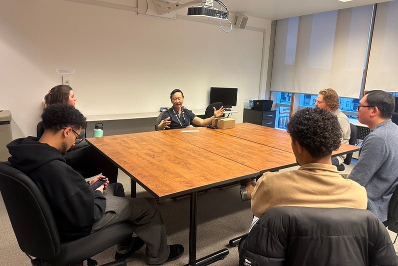 Small group of adults seated around a large rectangular wooden table in a classroom or meeting room. One person seated at the far side of the table speaks while gesturing with both hands, while others sit in rolling office chairs facing inward. The room has light-colored walls, a ceiling-mounted projector, and windows with partially lowered blinds. Personal items such as jackets, notebooks, and a takeaway cup are visible, suggesting a facilitated discussion or small-group meeting.