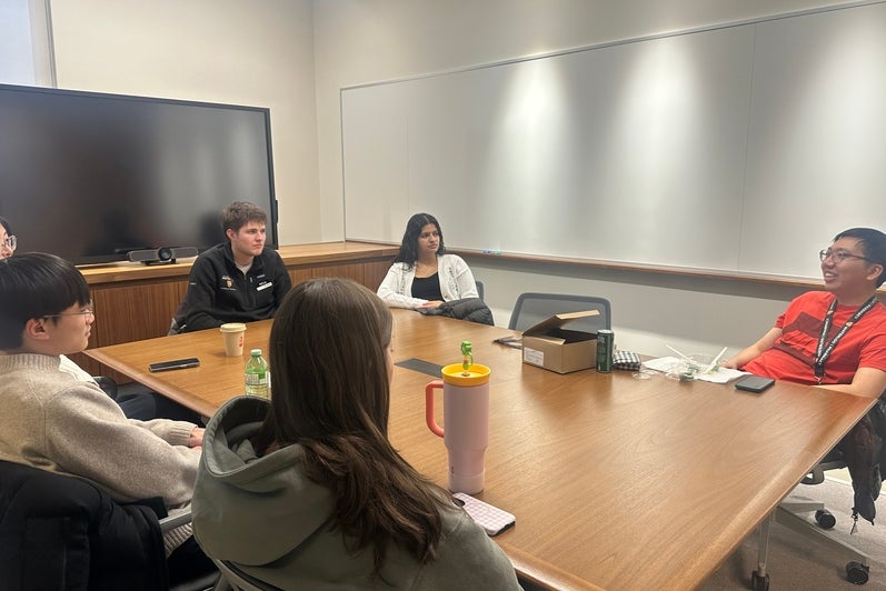 Small group of adults seated around a rectangular wooden conference table in a meeting room. Participants sit in office chairs facing inward, engaged in a discussion, with one person speaking near the far end of the table. Items such as notebooks, reusable water bottles, coffee cups, and a small box are placed on the table. The room features light-colored walls, a large wall‑mounted display screen, and bright overhead lighting, indicating a collaborative meeting or discussion setting.