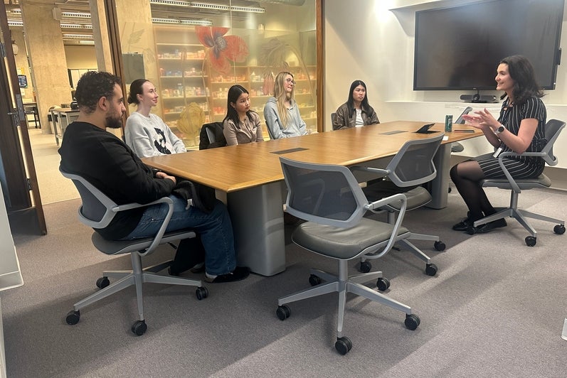 Small group of adults seated around a rectangular wooden meeting table in a conference room. One person speaks while gesturing, and others sit in rolling office chairs facing the speaker. A large wall-mounted display screen, glass walls, and a visible hallway or open workspace appear in the background. Personal items, including notebooks and a drink can, sit on the table, indicating a collaborative meeting or discussion setting.