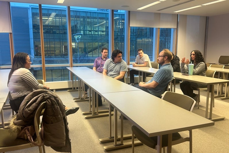 Medium-sized classroom with rows of rectangular tables facing a seated speaker at the front of the room. Several adults sit at the tables, oriented toward the speaker, who is seated on a stool near a podium with a computer monitor. The room features neutral-colored walls, overhead lighting, an exposed ceiling with ducts, a wall clock, and windows along one side. Personal items, including a reusable drink can on the floor, are visible, suggesting a lecture, talk, or facilitated discussion.