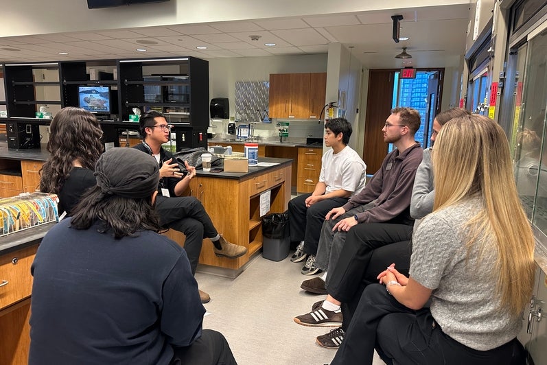 Small group of adults seated in a semicircle inside a laboratory or teaching lab, participating in a discussion. One person gestures while speaking near a central lab bench with cabinets, equipment, and supplies, while others sit on stools along the walls, wearing casual clothing and lanyards under bright indoor lighting.