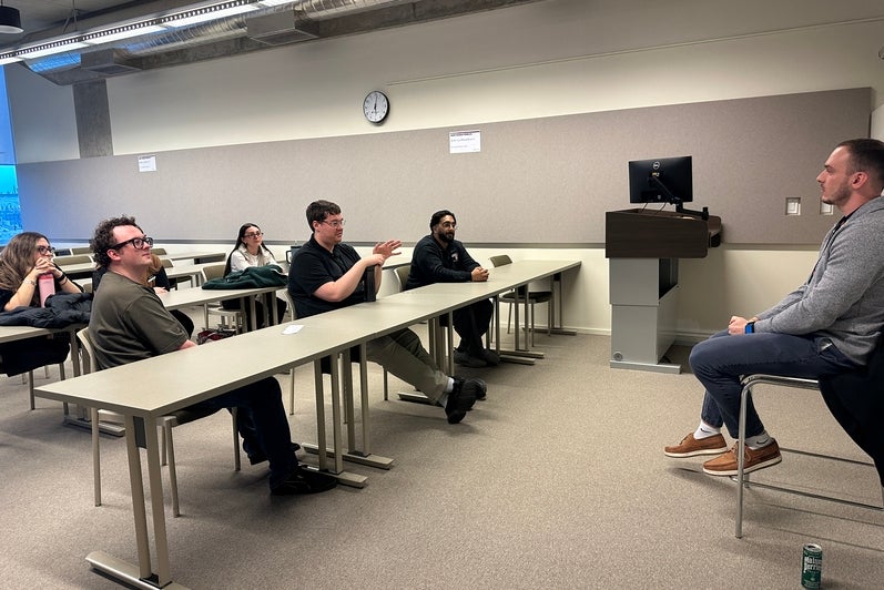 Medium-sized classroom with rows of rectangular tables facing a seated speaker at the front of the room. Several adults sit at the tables, oriented toward the speaker, who is seated on a stool near a podium with a computer monitor. The room features neutral-colored walls, overhead lighting, an exposed ceiling with ducts, a wall clock, and windows along one side. Personal items, including a reusable drink can on the floor, are visible, suggesting a lecture, talk, or facilitated discussion.