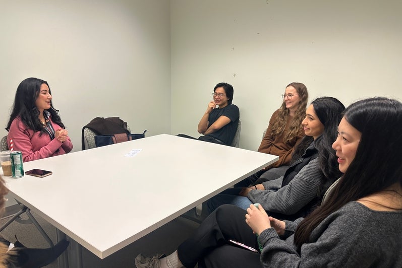 Small group of adults seated around a rectangular white table in a plain meeting room, engaged in conversation. One person speaks with hands clasped while others sit along the table edges, facing toward the speaker. Personal items such as notebooks, phones, a reusable cup, and outerwear are visible on and around the table. The room has light-colored walls, simple chairs, and even overhead lighting, suggesting an informal discussion or small-group meeting.