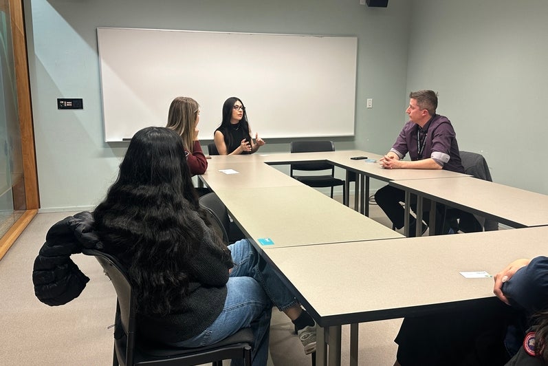 Small group of adults seated around rectangular tables arranged in a U-shape in a classroom or meeting room. One person at the far side of the tables appears to be speaking and gesturing, while others sit and listen. The room has light-colored walls, a whiteboard mounted at the front, and simple chairs. Personal items such as notebooks and jackets are visible, and the space is evenly lit, suggesting a discussion-based meeting or workshop.