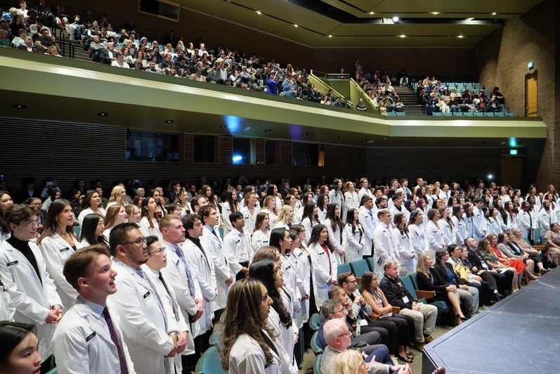 Group of pharmacy students in white coats standing in an auditorium during a ceremony, with audience members seated in front.