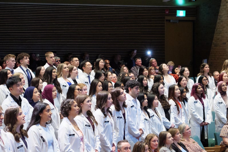 Pharmacy students in white coats standing together during a ceremony in an auditorium.