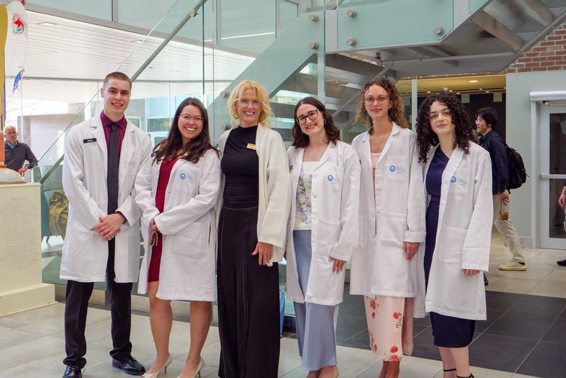 Small group of pharmacy students in white coats posing with a faculty member in a bright campus atrium.