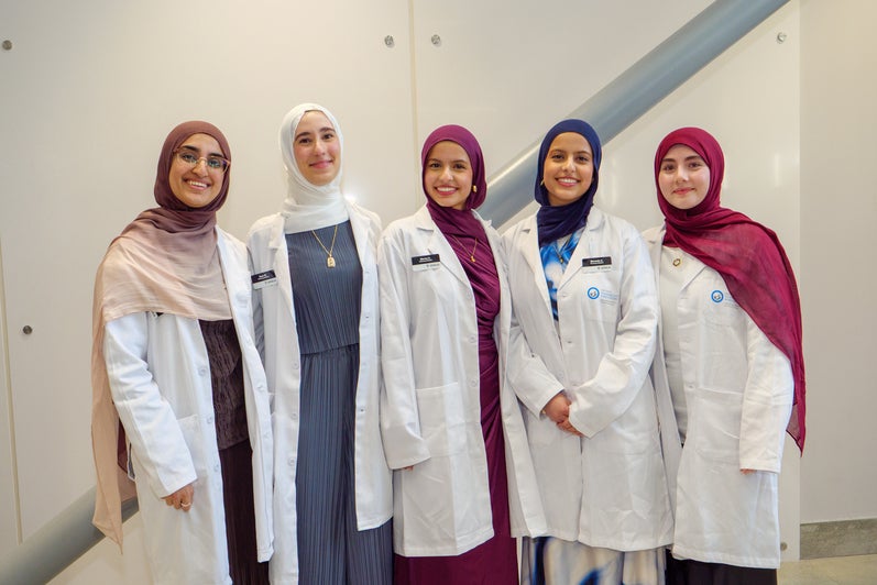 Five pharmacy students in white coats and headscarves posing together indoors.