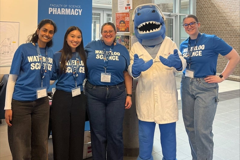 Five people wearing blue “Waterloo Science” shirts stand smiling beside a blue shark mascot in a white lab coat at a Faculty of Science Pharmacy booth.