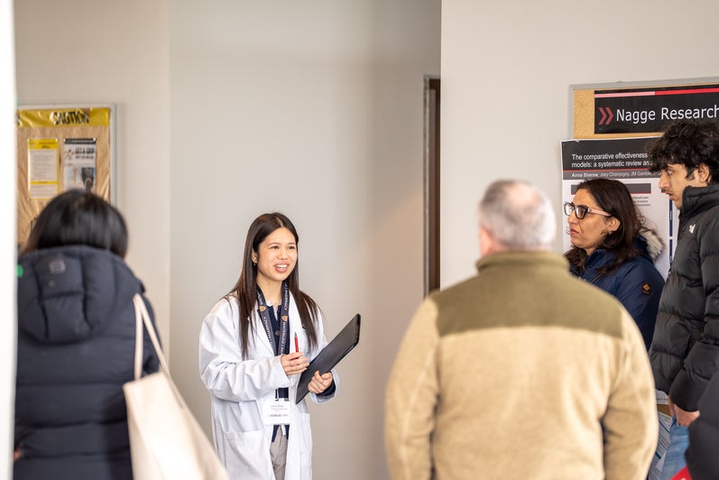 A person wearing a white lab coat speaks with a visitor while holding a folder in a campus hallway.