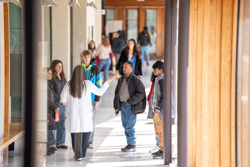 A person wearing a white lab coat speaks to a small group of people standing in a bright campus hallway.