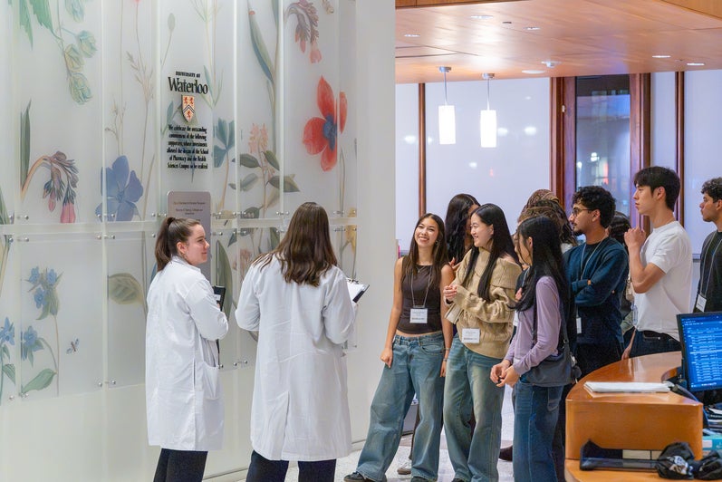 A group of attendees wearing name badges gather near two people in white lab coats in a University of Waterloo building, with a floral wall display and University of Waterloo crest in the background.