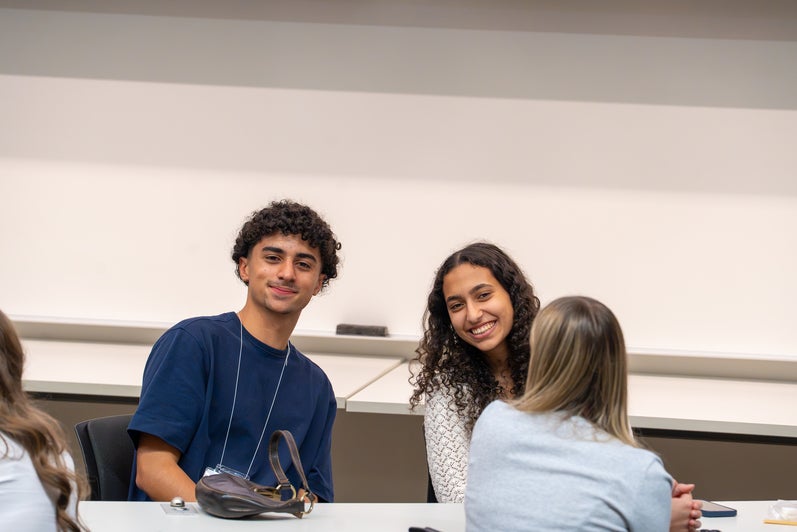 Three attendees sit together in a classroom during an event, with two facing the camera and another seated across from them at a desk.