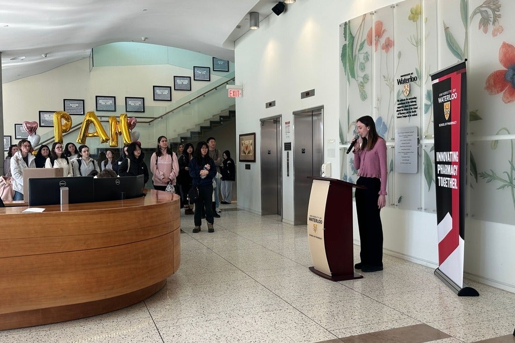 A person stands at a podium with a microphone in a bright building lobby decorated with large floral wall art. A University of Waterloo banner reading “Innovating Pharmacy Together” is positioned beside the podium. In the background, a group of people stands near a staircase, facing the speaker. Gold balloon letters forming “PAM” and two heart-shaped balloons are displayed behind the group near the reception desk. Two elevators and wall-mounted plaques are visible along the back wall.