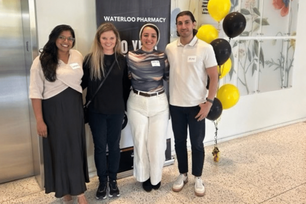 Four people stand together at a Waterloo Pharmacy event beside a Waterloo Pharmacy banner and a cluster of black and yellow balloons.