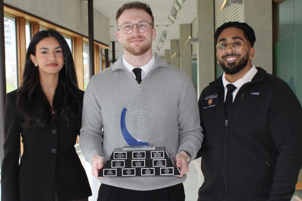Three students pose in a campus hallway, with one holding a multi‑plate award trophy.
