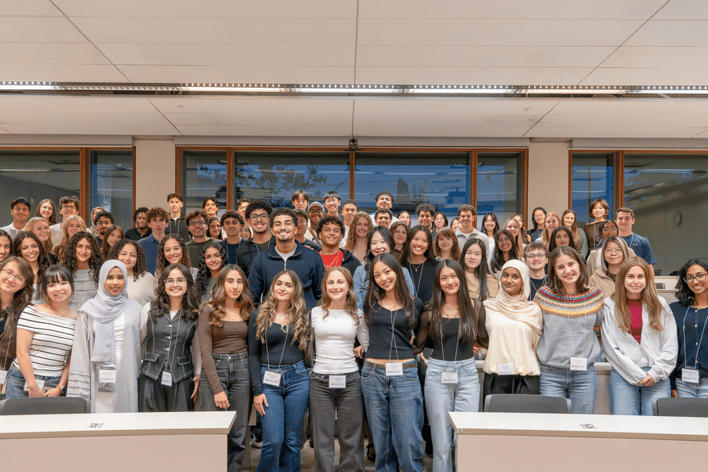 A large group of students pose together for a group photo in a bright classroom, arranged in several rows between desks and wearing name badges.