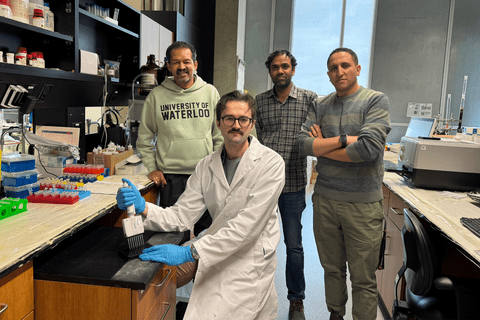 Four people are positioned in a laboratory setting with benches, instruments, and lab supplies visible. One person in the foreground sits at a lab bench wearing a white lab coat and blue gloves while holding a pipette over laboratory equipment; three others stand behind with arms relaxed or crossed. Shelves, bottles, tubes, and devices surround the workspace, and a sweatshirt in the background displays the text “University of Waterloo.”