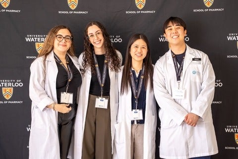 Four people wearing white lab coats stand smiling in front of a University of Waterloo School of Pharmacy backdrop, each wearing a conference name badge.