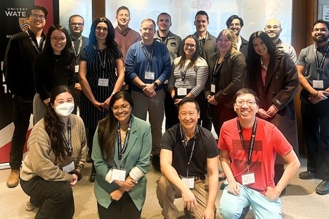 Group photo of approximately 18 adults wearing conference lanyards, arranged in two rows inside a modern indoor meeting space. The individuals are dressed in business-casual attire and stand or kneel in front of a light-colored wall with partial signage visible, including University of Waterloo branding and a sign that reads “Quiet Please Meeting in Progress.” Lighting is bright and even, and the image appears to document a professional gathering or workshop.