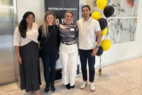 Four people stand together at a Waterloo Pharmacy event beside a Waterloo Pharmacy banner and a cluster of black and yellow balloons.