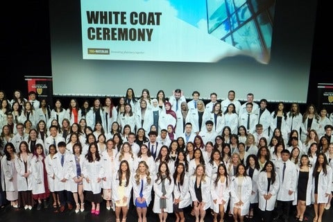 Large group of University of Waterloo pharmacy students in white coats posed on stage under a “White Coat Ceremony” banner, with school banners on either side.