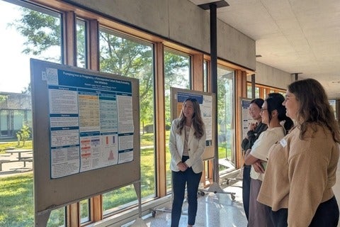 A presenter discusses research findings beside a poster display as several attendees listen during a poster session. The event takes place in a bright corridor with floor-to-ceiling windows, natural light, and multiple research posters mounted on display boards.