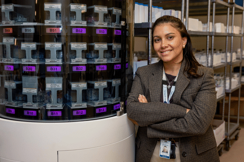 Person standing beside an automated medication dispensing system in a hospital pharmacy.