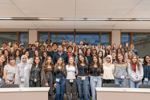 A large group of students pose together for a group photo in a bright classroom, arranged in several rows between desks and wearing name badges.