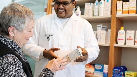 A male pharmacist holding a box of medicine and speaking to an elderly female patient.