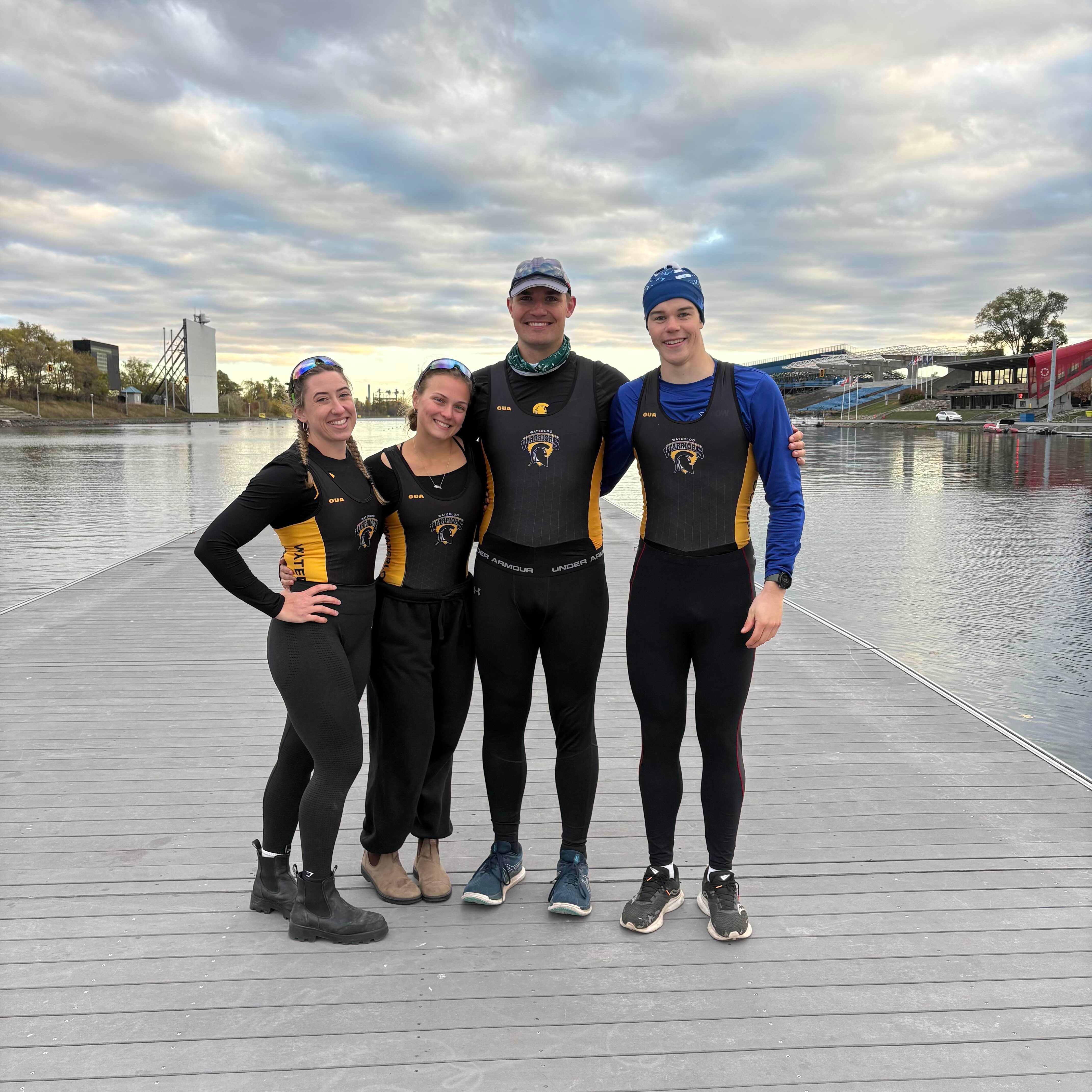 Four rowing team members stand together on a wooden dock beside a calm watercourse under a cloudy sky. All are wearing black and gold Waterloo Warriors rowing uniforms, with one person in a blue long-sleeve layer and another wearing a cap. The group stands close together with arms around each other. In the background, there is a rowing basin with lanes, a starting tower, and grandstand seating along the shoreline.